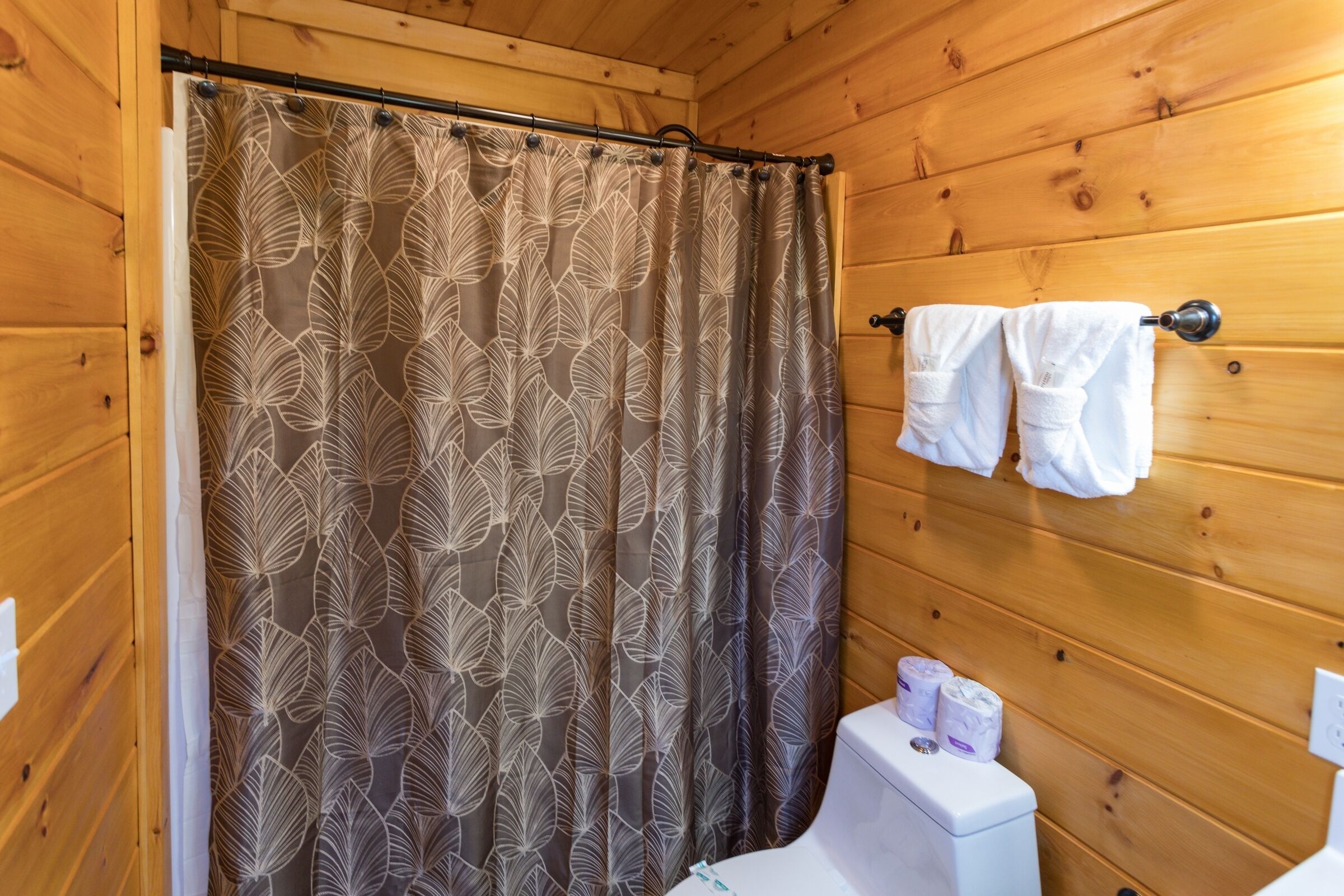 Bany d'una cabana de fusta amb una cortina de bany amb dibuixos de fulles, un vàter amb paper higiènic i tovalloles penjades en un suport.