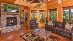 A spacious living room with a stone fireplace, leather furniture, and a large window overlooking a forested area. - Luxurious 5-Bedroom Cabin with Indoor Pool (SEVIERVILLE)