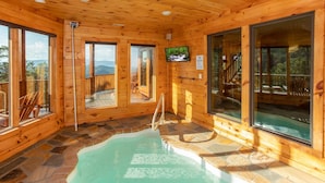A wooden cabin with a stone-tiled pool, large windows, and a television. - Luxurious 5-Bedroom Cabin with Indoor Pool (SEVIERVILLE)