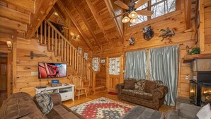 A cozy log cabin interior with a wooden staircase, a fireplace, and a television in the living room. - Cozy Den Cabin with Covered Porch and Games (PIGEON FORGE)