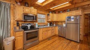 A wooden cabin kitchen with a stainless steel stove, microwave, and refrigerator, wooden cabinets, and a window overlooking the outside. - Cozy Den Cabin with Covered Porch and Games (PIGEON FORGE)
