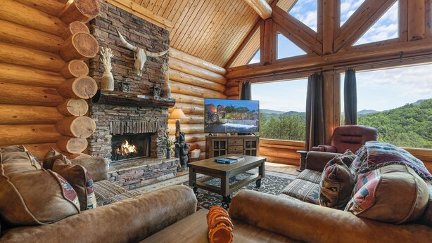 L’intérieur d’une cabane en rondins, avec une cheminée en pierre, des troncs d’arbres apparents, un téléviseur et une vue sur l’extérieur.