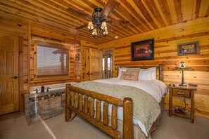 A wooden cabin bedroom with a bed, a nightstand, a window with blinds, a ceiling fan, and a framed picture on the wall.