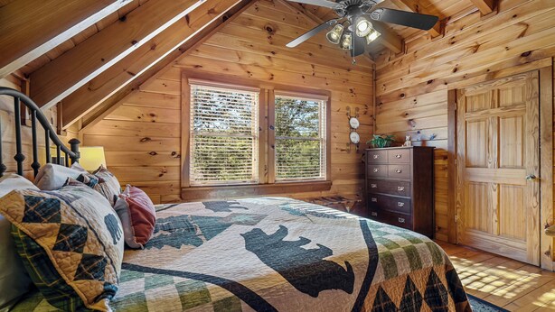 Ein gemütliches Schlafzimmer mit Holzbalkendecke, einem Bett mit Steppdecke, einem Schminktisch und einem Fenster mit Jalousien.