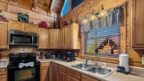 A wooden cabin kitchen with a microwave, stove, sink, and window with blinds.