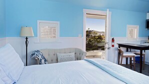 A bedroom with a bed, a desk, and a chair. There is a window with blinds, a lamp, and a fire extinguisher.