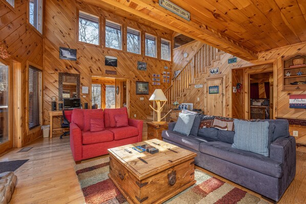 A cabin interior with wooden walls, a red and blue sofa, a wooden coffee table, and a staircase. - Cozy Cabin with Fireplace (SEVIERVILLE)