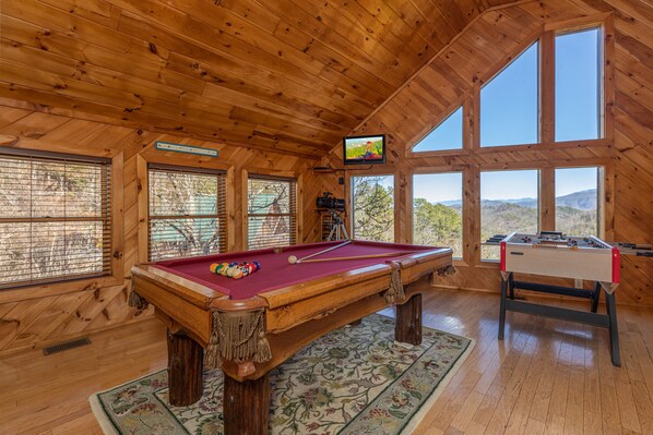 Um quarto com piso de madeira, mesa de bilhar, mesa de pebolim e amplas janelas com vista para o exterior.