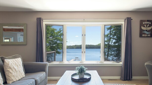 A living room with a large window overlooking a body of water, a gray sofa, a coffee table with a plant, and a sign on the wall. - Dollar Lake Oasis, Near Downtown and the Chain, Fire Pit, Sandy Beach, W&D, WiFi (Eagle River)