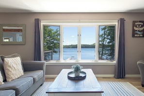 A living room with a large window overlooking a body of water, a gray sofa, a coffee table with a plant, and a sign on the wall.