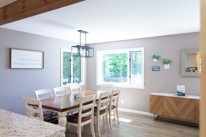 A dining area with a wooden table and chairs, a chandelier, and a view of greenery through the windows.