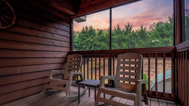 Una terraza de madera con dos mecedoras y una mesita, con vistas a un área boscosa.