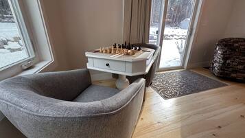 Une pièce chaleureuse avec un fauteuil gris, une petite table d’appoint blanche surmontée d’un jeu d’échecs, et un tapis à motifs posé sur un plancher en bois.