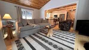 A living room with a sofa, coffee table, and patterned rug. There is a kitchen visible through an open doorway.