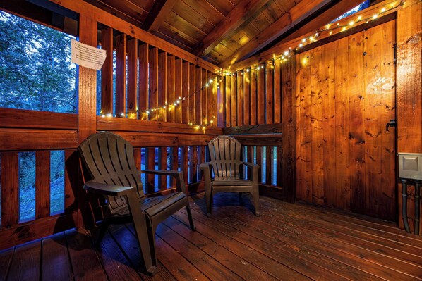 A wooden deck with Adirondack chairs, string lights, and a view of trees through a window. - Gatlinburg Celebration Destination (GATLINBURG)