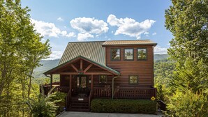 A two-story log cabin with a porch and a covered front entrance, surrounded by lush greenery and mountains in the background. - Galloping In The Mountains (PIGEON FORGE)