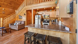 A spacious cabin with a kitchen area, a dining area with a couch and stools, and a wooden staircase. - Galloping In The Mountains (PIGEON FORGE)