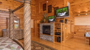Interno di una baita in legno con camino, due televisori, uno specchio e un letto con copripiumino decorato.