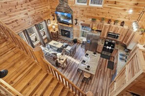 Interior de uma cabana de madeira com sala de estar, cozinha, lareira e grandes janelas.