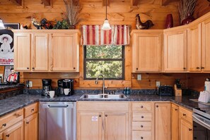 A kitchen with wooden cabinets, a stainless steel dishwasher, a coffee maker, and a window with a view of trees.