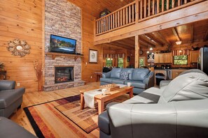A cozy cabin interior with a stone fireplace, a television mounted on the wall, a dining area with a table and chairs, and a balcony with wooden railings.