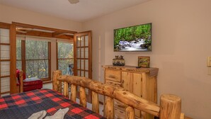 A bedroom with a wooden bed, a television mounted on the wall, and a view of the outdoors through large windows. - Pigeon Forge*Easy Access Rds*WiFi*Hot Tub (SEVIERVILLE)