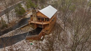 A large, multi-story log cabin with a wrap-around deck and a stone chimney, surrounded by a wooded area. - Pigeon Forge*Easy Access Rds*WiFi*Hot Tub (SEVIERVILLE)