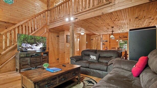 Ein Holzhütten-Interieur mit einer Holztreppe, einem Ledersofa, einem Fernseher auf einem Holzkabinett und einem Essbereich im Hintergrund.