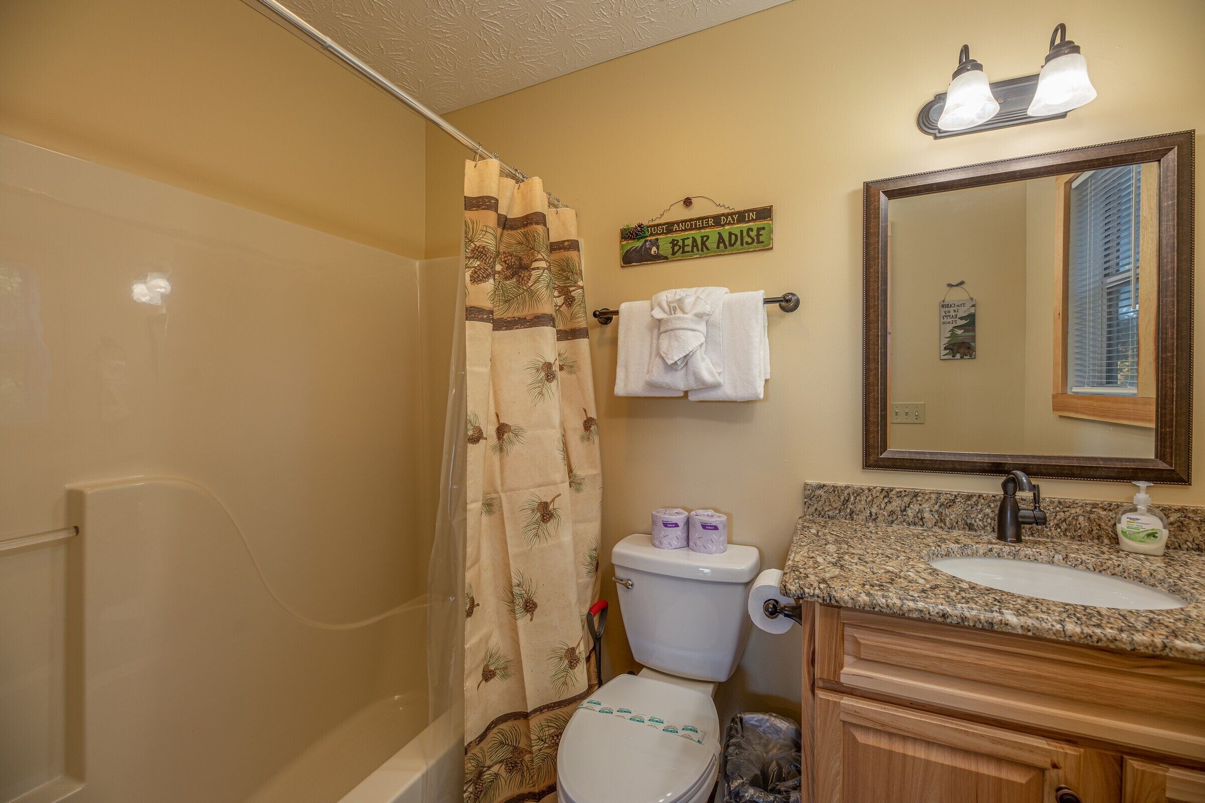 A bathroom with a shower curtain, toilet paper, a towel rack, a sink with a mirror, and a sign above the toilet.