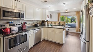 A kitchen with white cabinets, stainless steel appliances, a red kettle, and a window with a view of greenery.