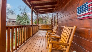 A wooden porch with a railing, wooden chairs, and a flag on the wall. - Simplicity Like It Should Be: In A Cabin (PIGEON FORGE)
