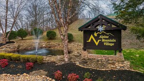 A sign for Alpine Mountain Village with a small pond and fountain, surrounded by landscaped bushes and trees. - Simplicity Like It Should Be: In A Cabin (PIGEON FORGE)