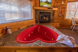 A wooden cabin interior with a stone fireplace, a red hot tub, and a television mounted above the fireplace.
