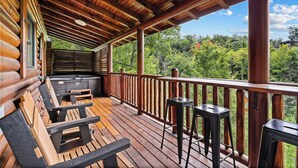 A wooden deck with seating and a hot tub, overlooking a forested area. - Filling Your Vacation Time With Fun And Games (PIGEON FORGE)