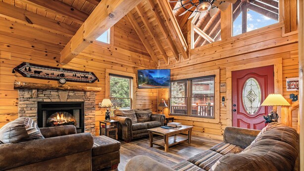 A cozy log cabin interior with a stone fireplace, leather sofas, wooden floors, and a large TV screen. - Filling Your Vacation Time With Fun And Games (PIGEON FORGE)