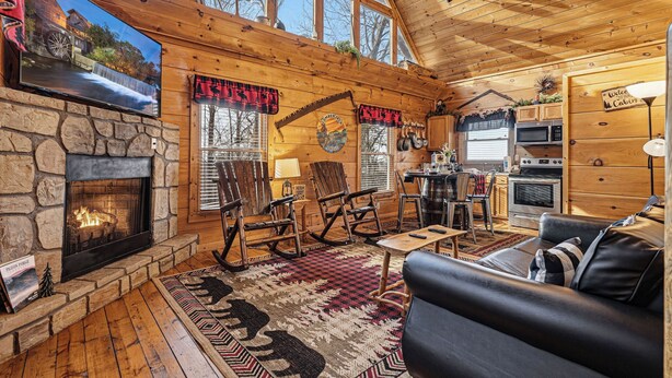 A cozy cabin interior with a stone fireplace, wooden walls, a television, rocking chairs, a sofa, and a kitchen area. - Relax And Enjoy The Incredible View From Here (GATLINBURG)