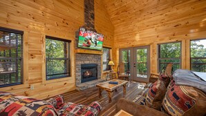 L’intérieur chaleureux d’un chalet avec une cheminée en pierre, des murs en bois et de grandes fenêtres donnant sur le paysage extérieur.