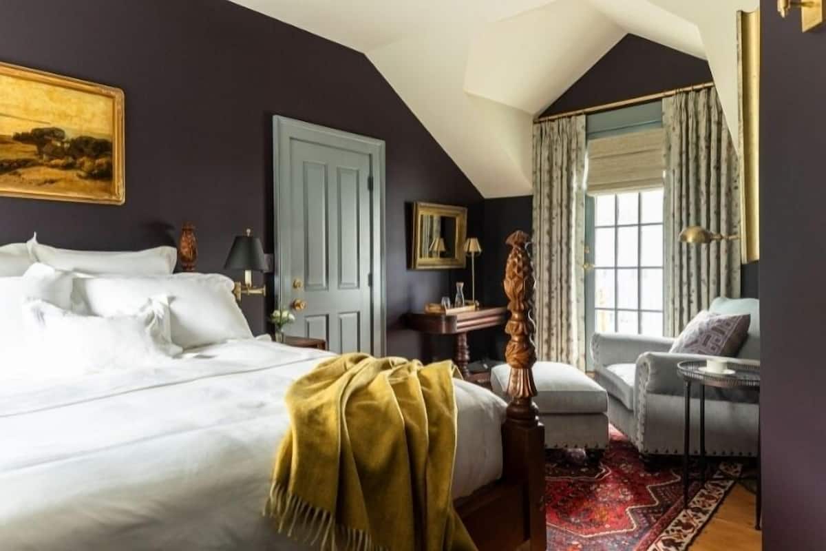 A bedroom with a large bed, a wooden chair, a small table, and a window with curtains.