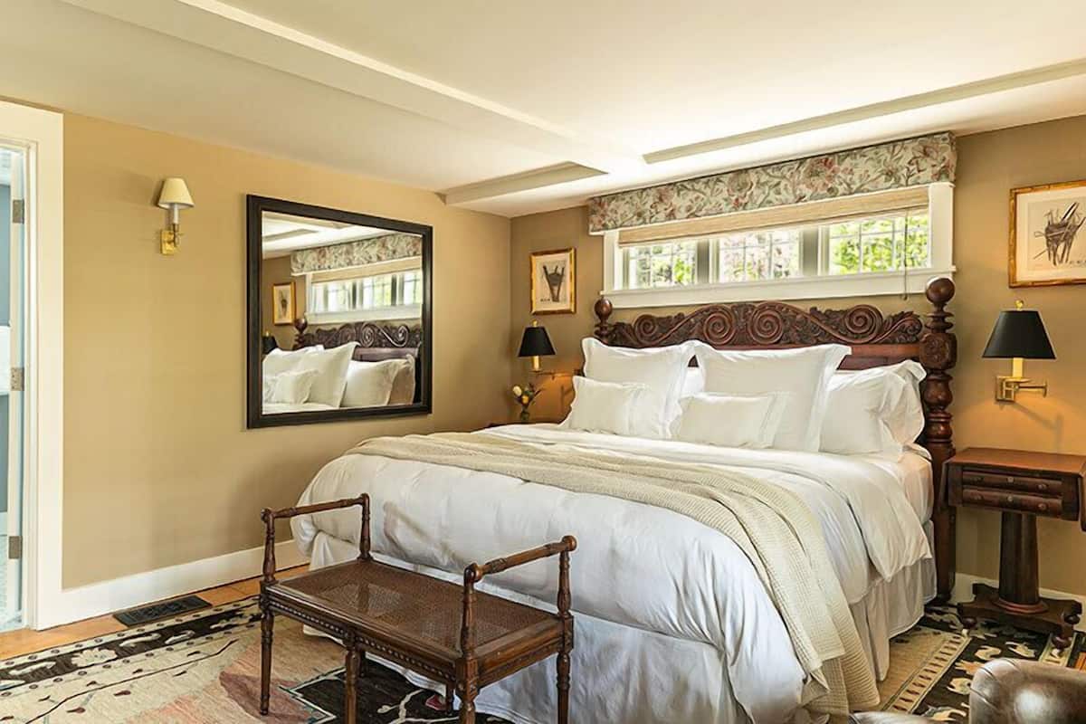 A bedroom with a large bed, a wooden chair, a mirror, and a window with a view.