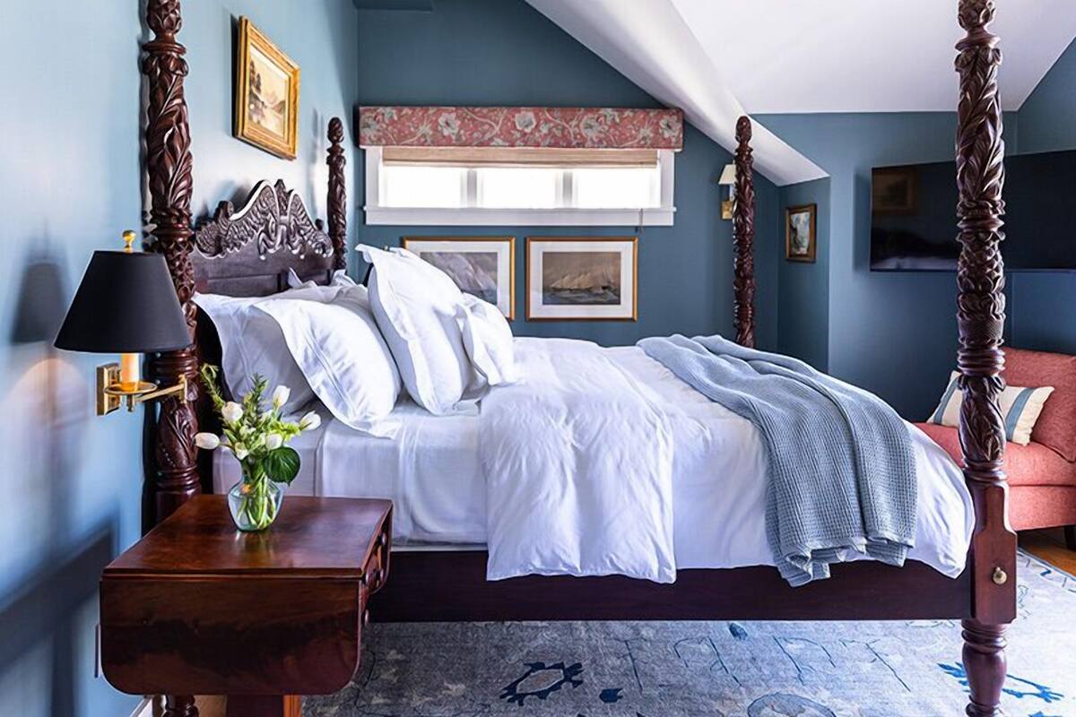 A four-poster bed with white linens, a vase of flowers, and a red sofa in a room with blue walls and a patterned rug.