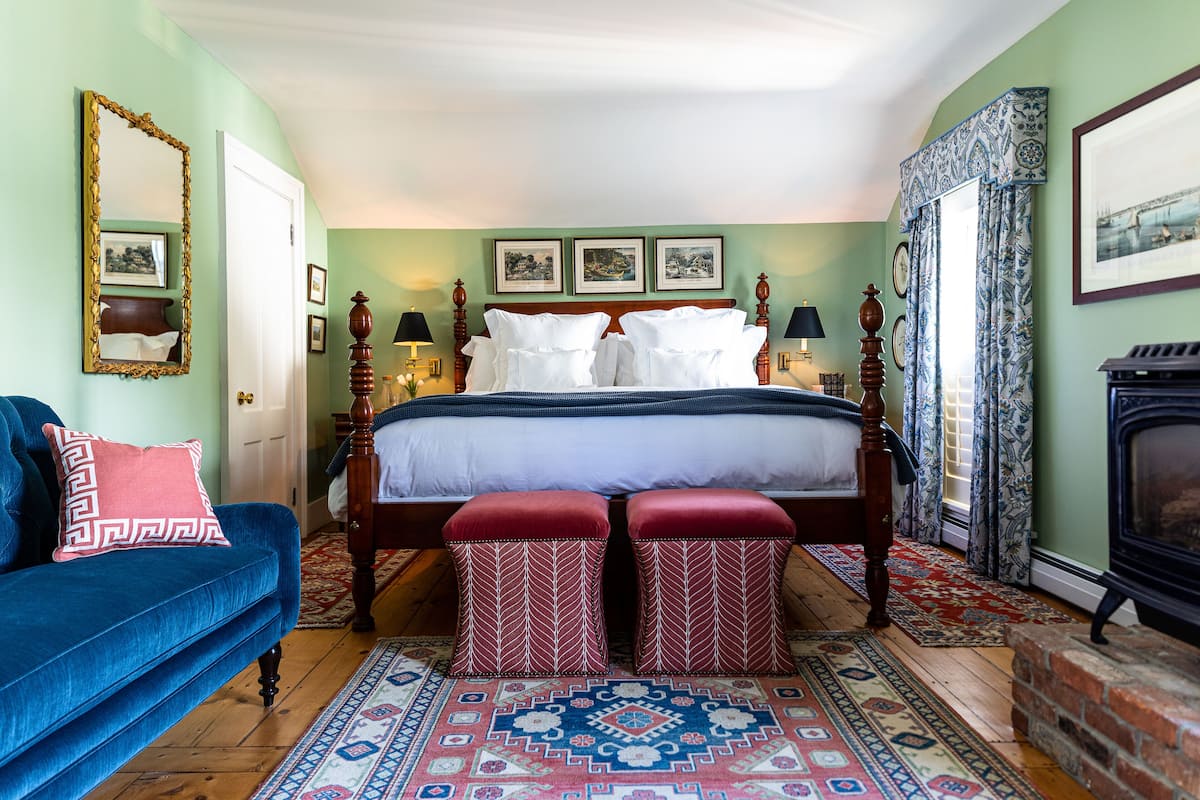A bedroom with a bed, a blue sofa, two red ottomans, a fireplace, and a patterned rug.