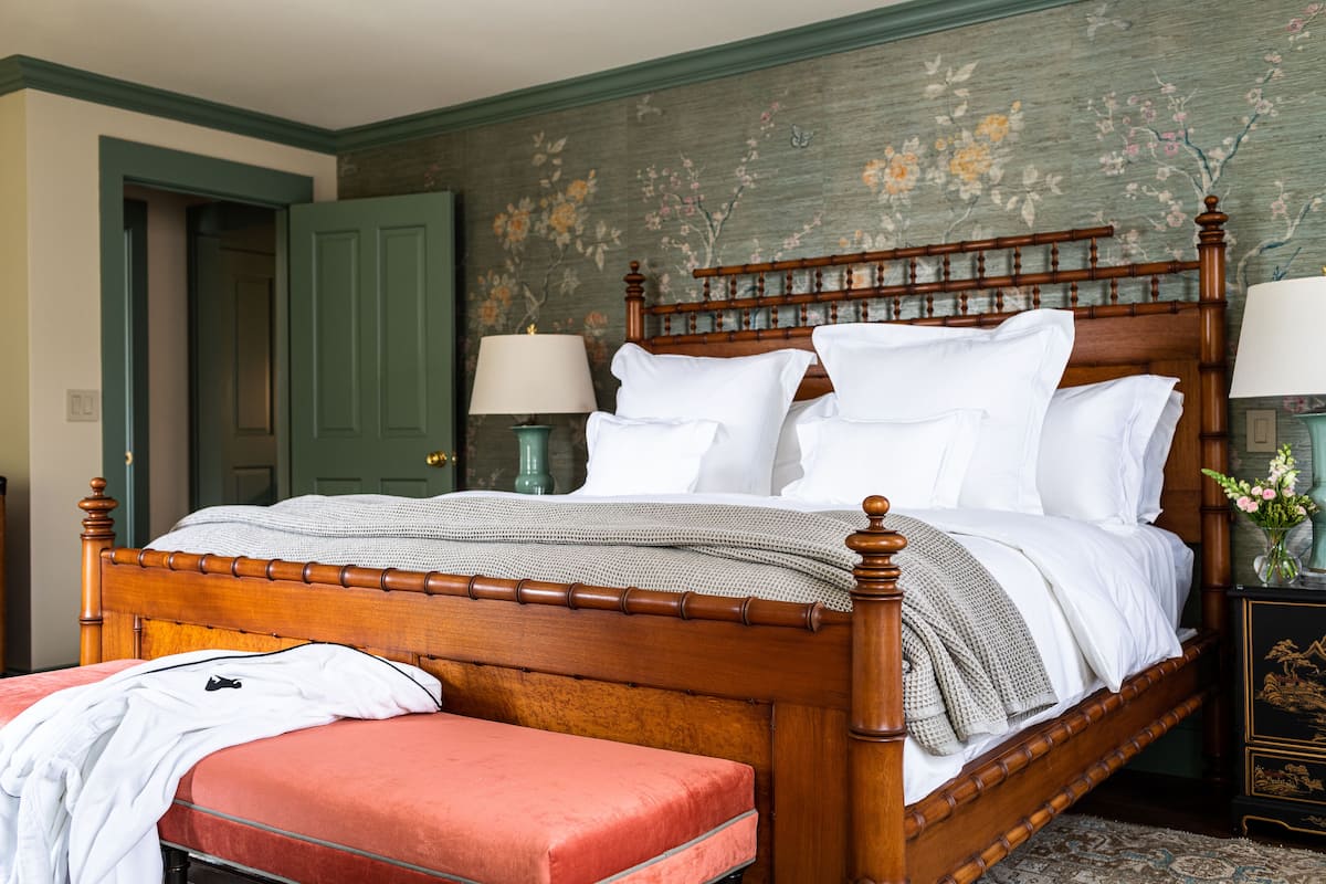 A neatly made bed with a wooden headboard, a red bench, and a nightstand with a lamp.