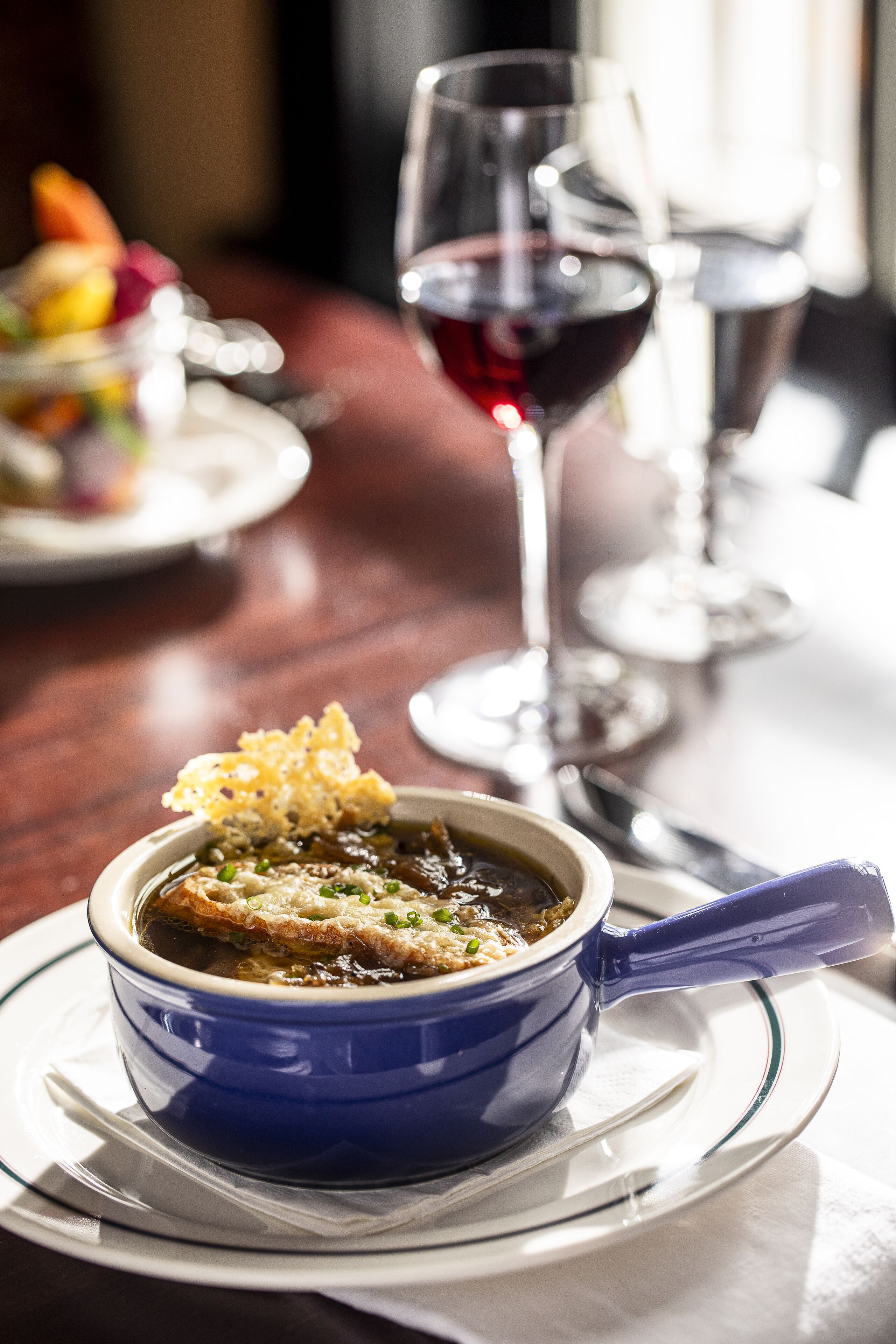 Mesa de jantar com uma tigela azul de sopa, um copo de vinho tinto e um prato de comida ao fundo.