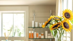 Un baño con repisa de vidrio con artículos de tocador, un florero con girasoles y una ventana con persianas.