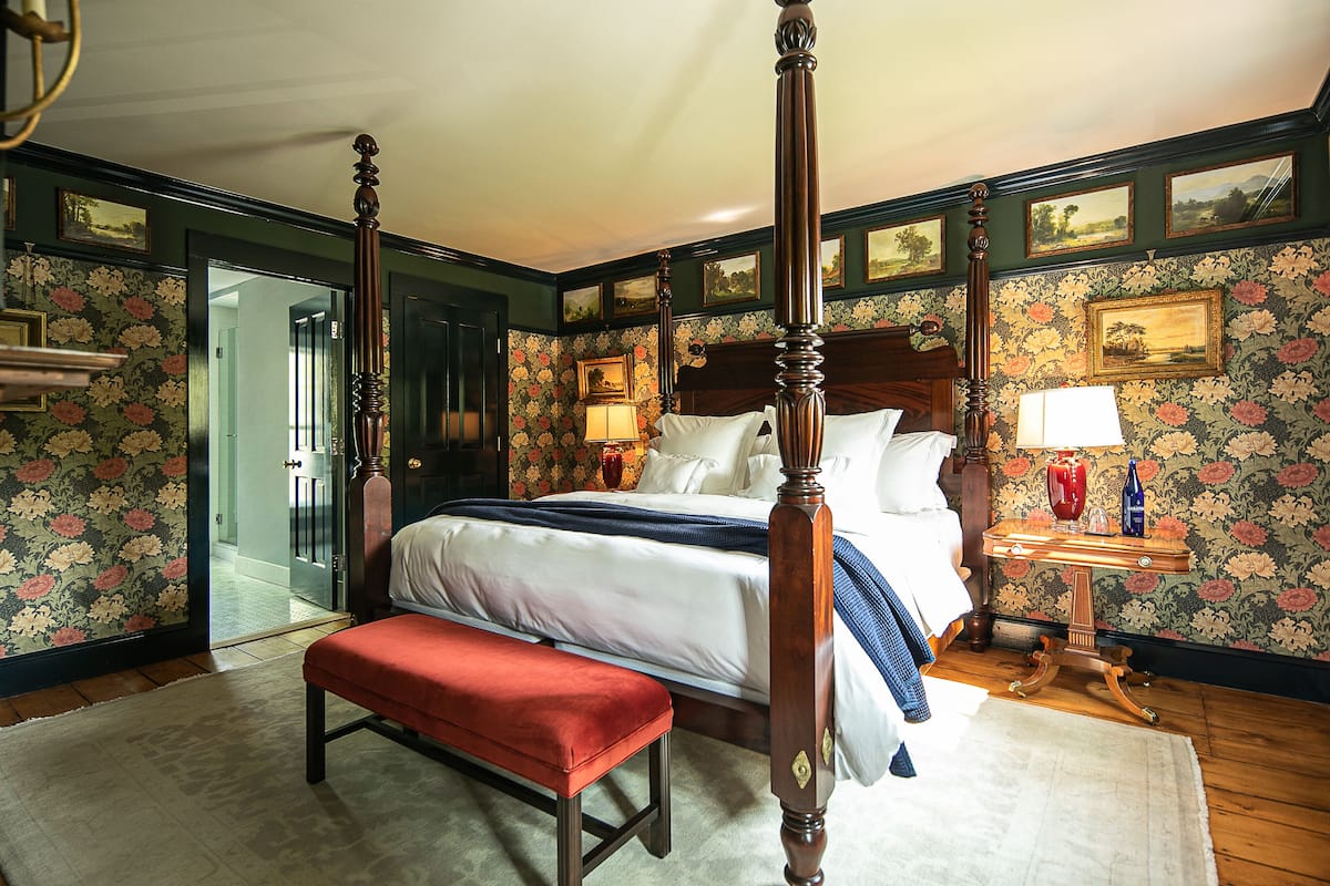 A four-poster bed with a wooden headboard, a bench, and a nightstand with a lamp.