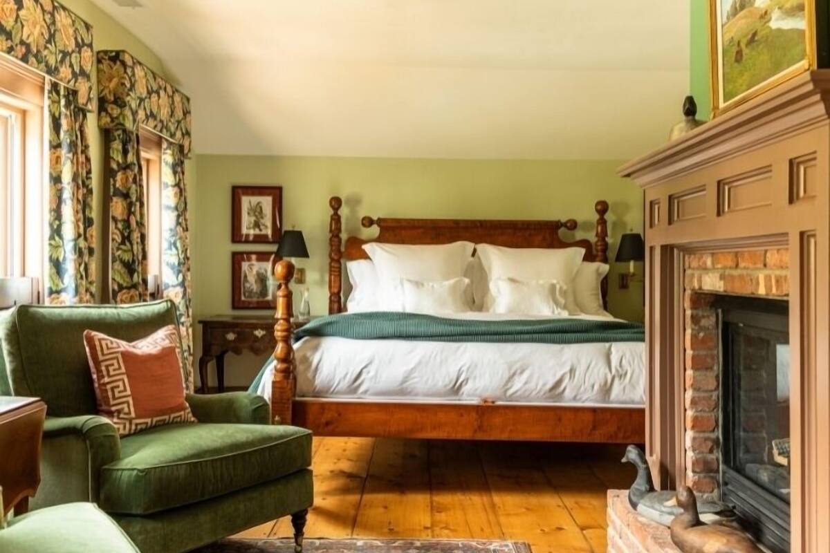 A traditional bedroom with a wooden bed, green armchair, and a fireplace.