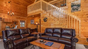 A cozy cabin interior with a wooden staircase, leather couches, a dining area, and a kitchen.