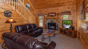 A log cabin interior with a fireplace, leather sofa, wooden staircase, and a television.