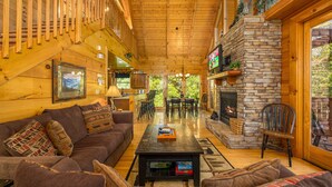Un intérieur de chalet chaleureux avec un plafond en bois, une cheminée en pierre, un ensemble de canapés et un coin repas.