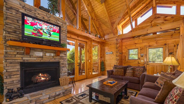 L’intérieur d’un chalet confortable, avec des murs en rondins de bois, une cheminée en pierre, un téléviseur fixé au mur et un coin salon accueillant, comprenant un canapé et une table basse.